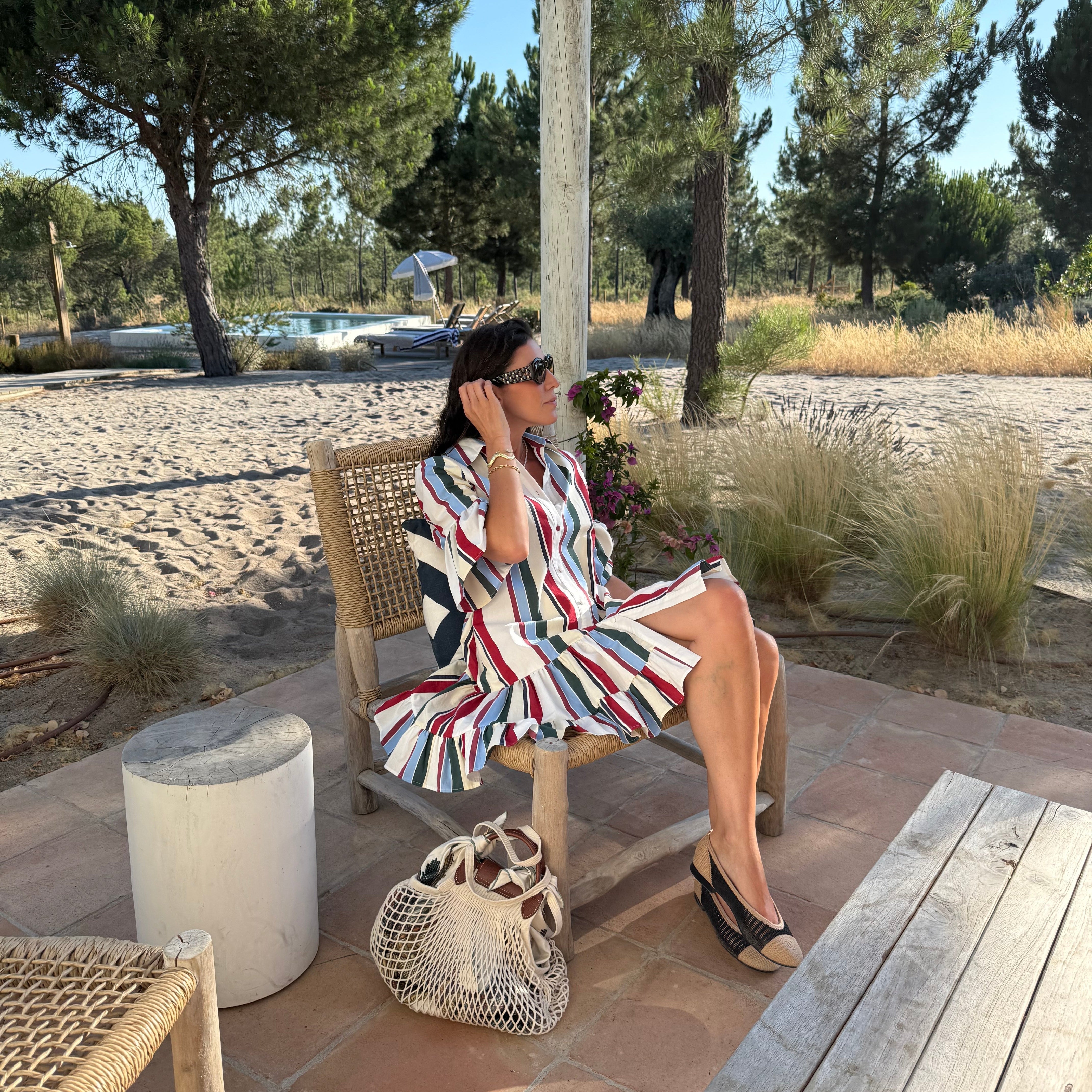 marianna wearing a stripy dress sitting on a rattan chair, outdoors in frot of a swimming pool
