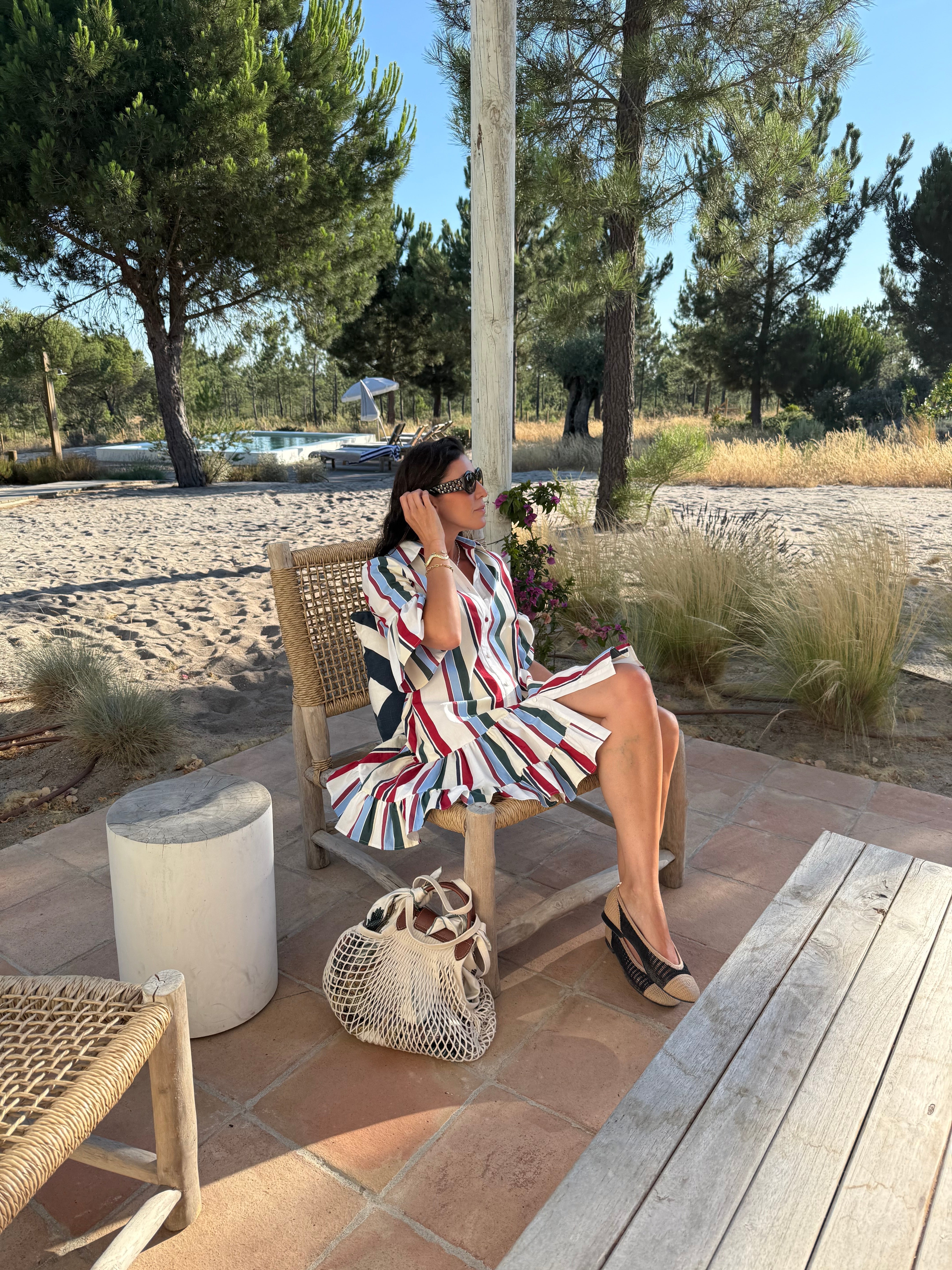 marianna wearing a stripy dress sitting on a rattan chair, outdoors in frot of a swimming pool