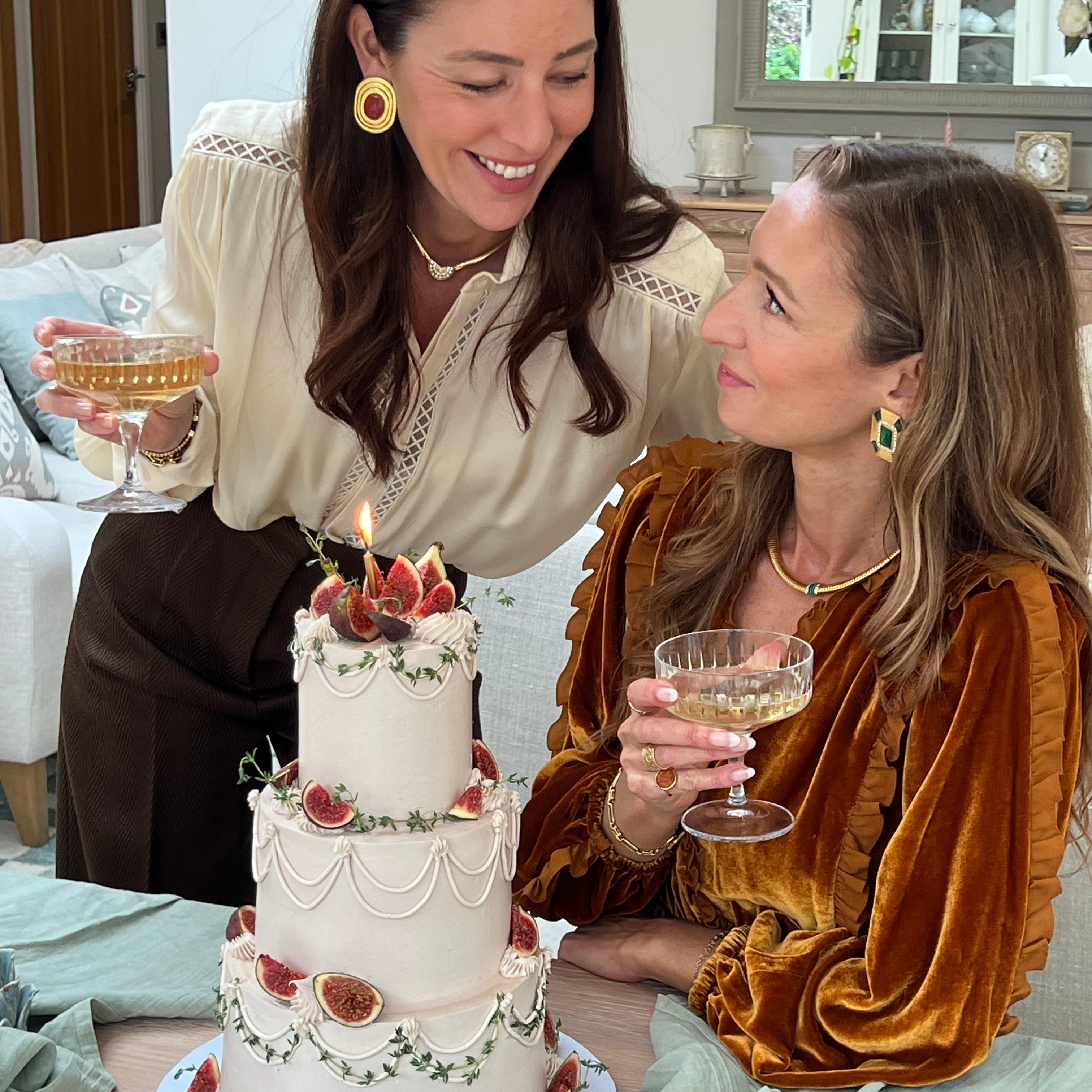 marianna and francesca from soru smiling next to a large 3 tired cake decorated with figs both holding a glass of champagne