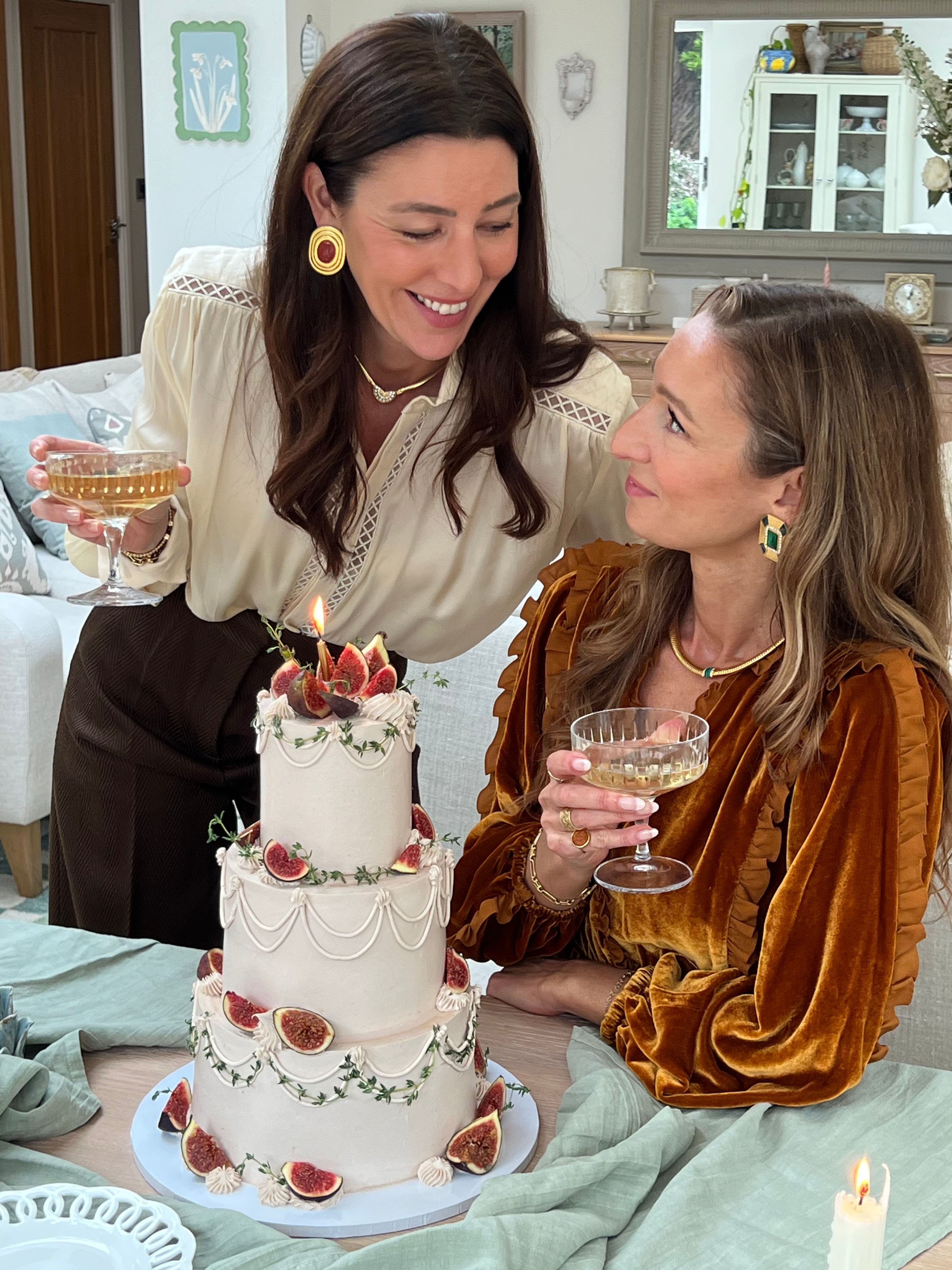 marianna and francesca from soru smiling next to a large 3 tired cake decorated with figs both holding a glass of champagne