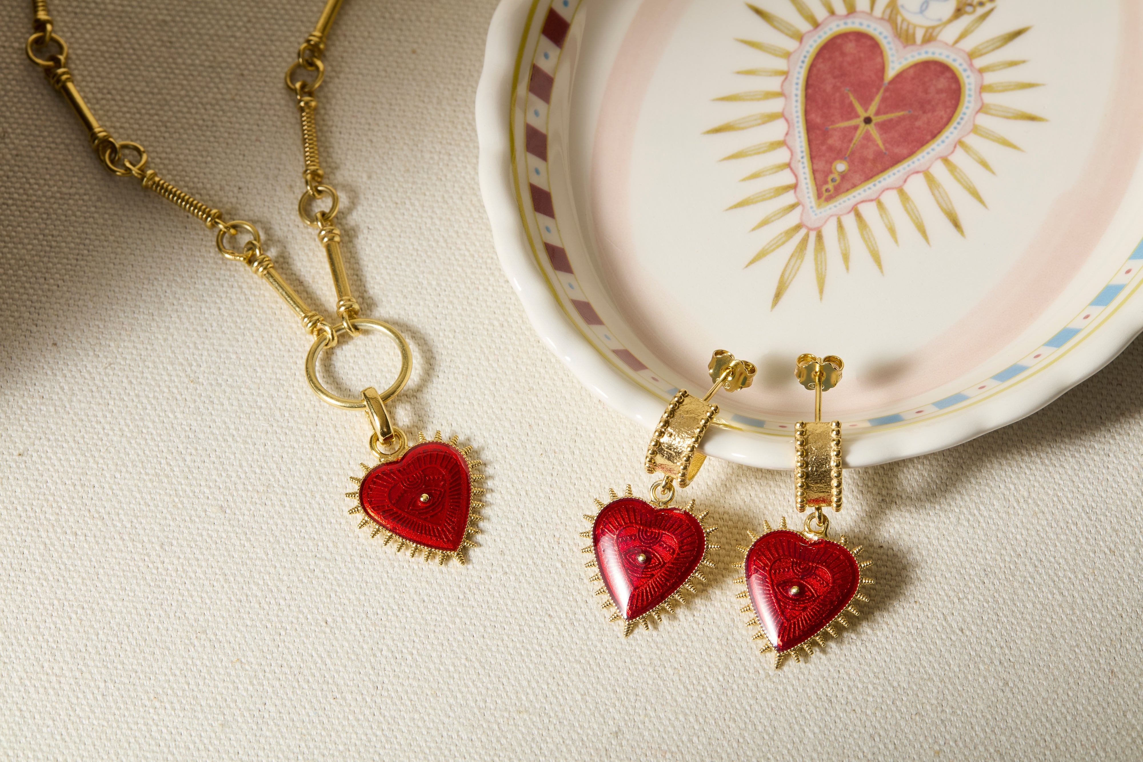 red enamel and gold heart earrings and charm necklace laid on a trinket dish with heart motif 