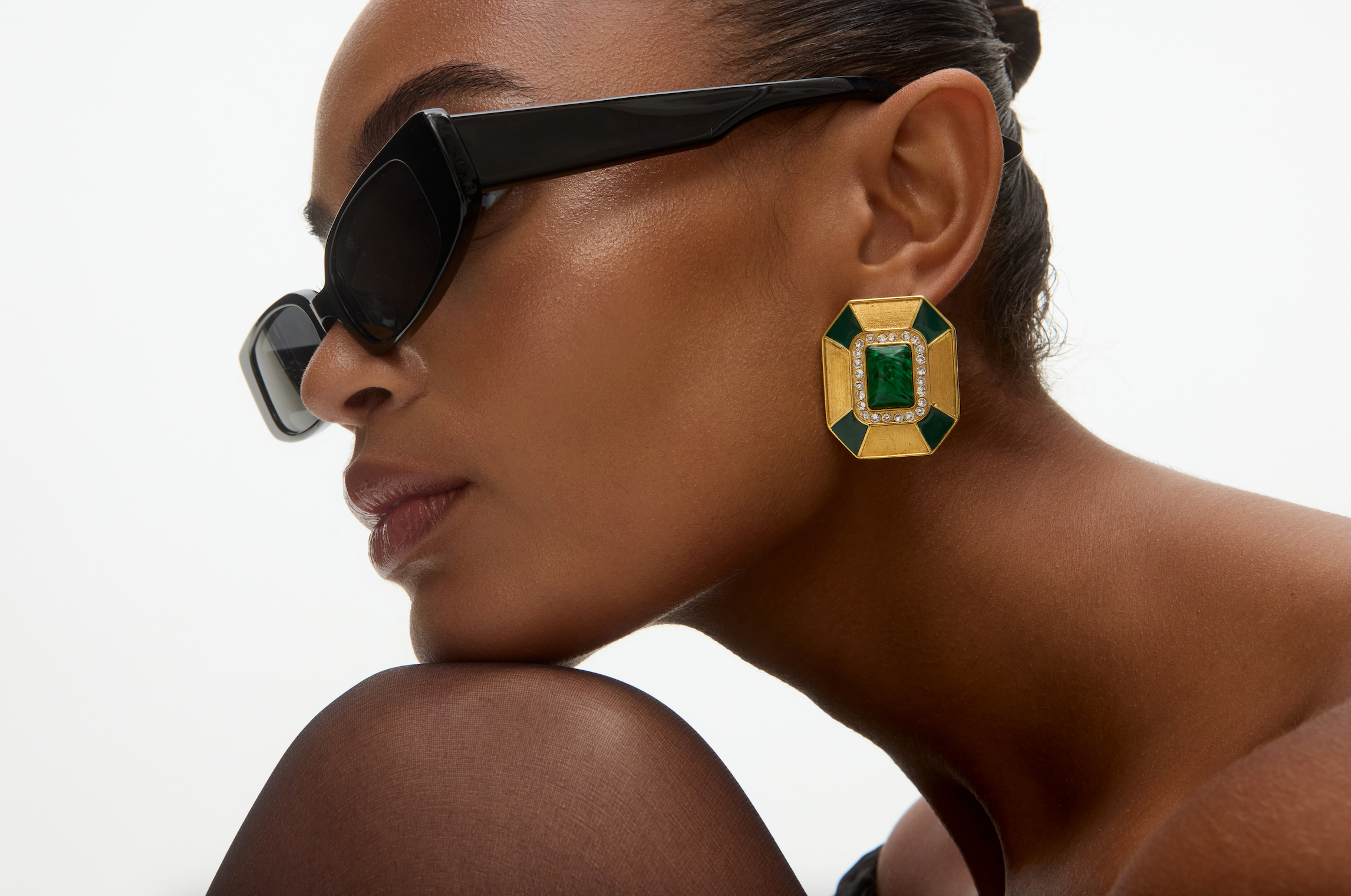 Model close up wearing a large green and gold art deco style earring with black sunglasses on a neutral background 