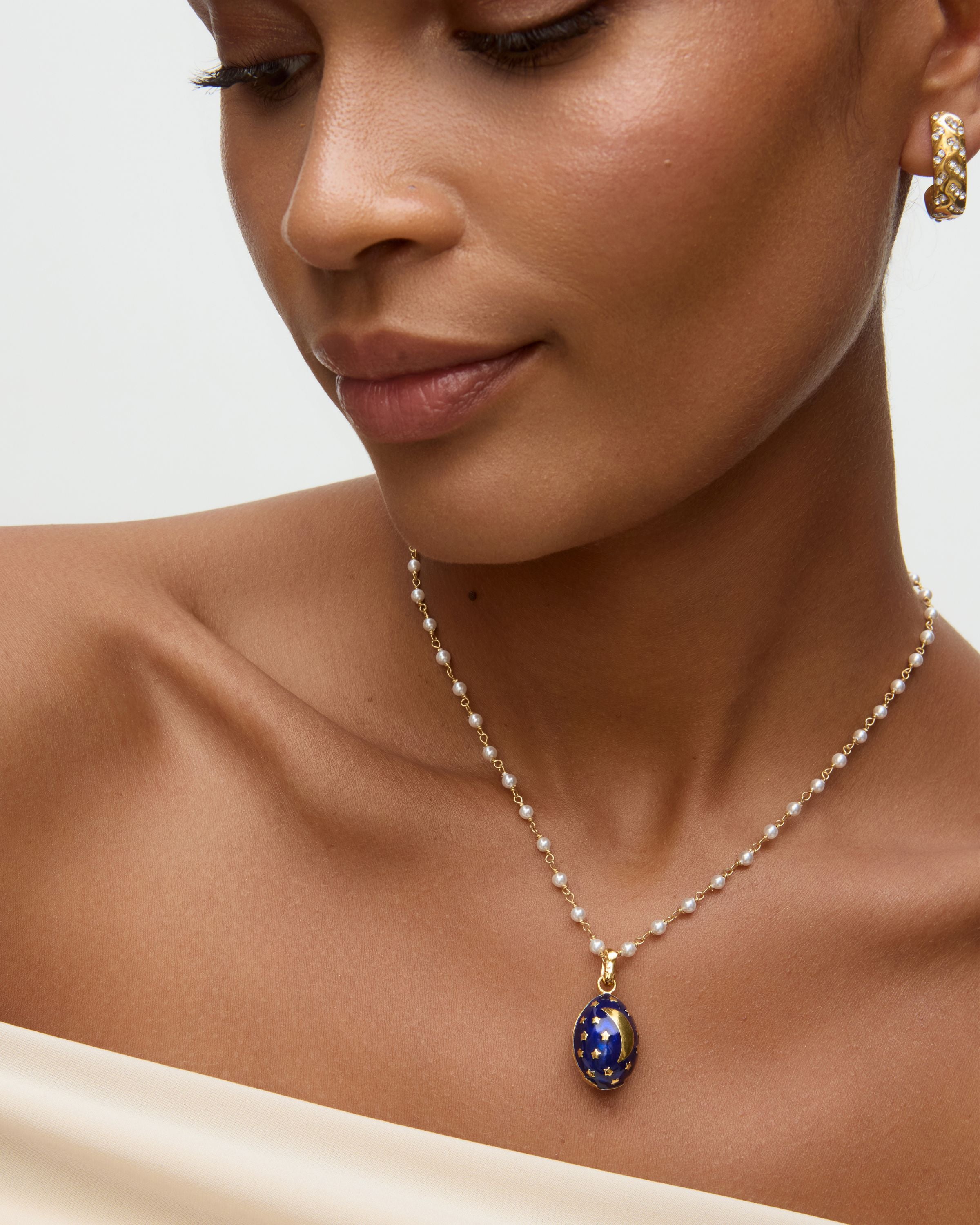 Close-up of a woman wearing a necklace with a blue egg shaped pendant with gold moon and stars on it  on a neutral background