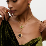 Woman wearing two layered gold charm necklaces with gemstones, green velvet top, and brown ring on a neutral background