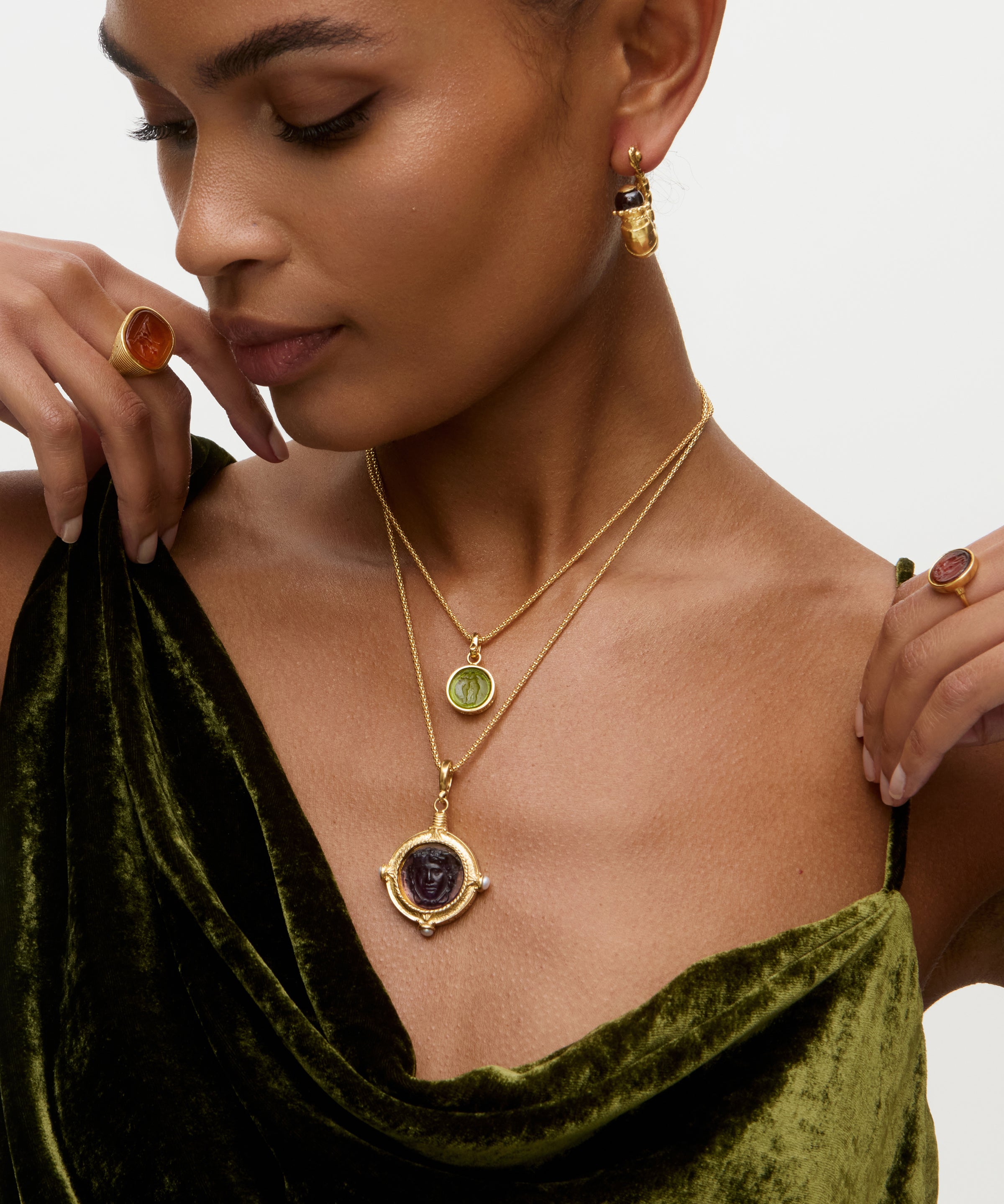 Woman wearing two layered gold charm necklaces with gemstones, green velvet top, and brown ring on a neutral background