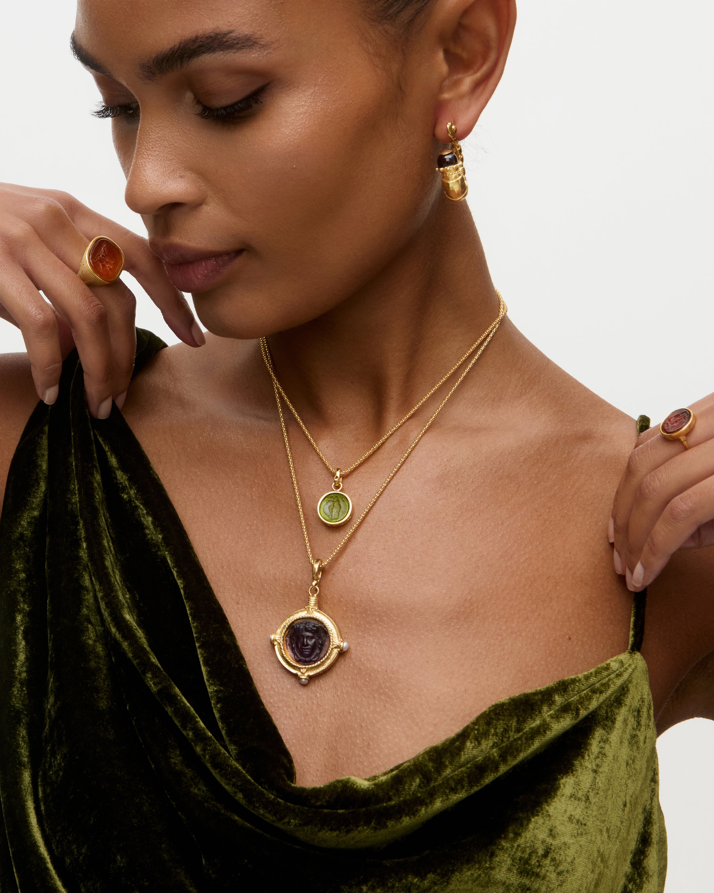 Woman wearing two layered gold charm necklaces with gemstones, green velvet top, and brown ring on a neutral background