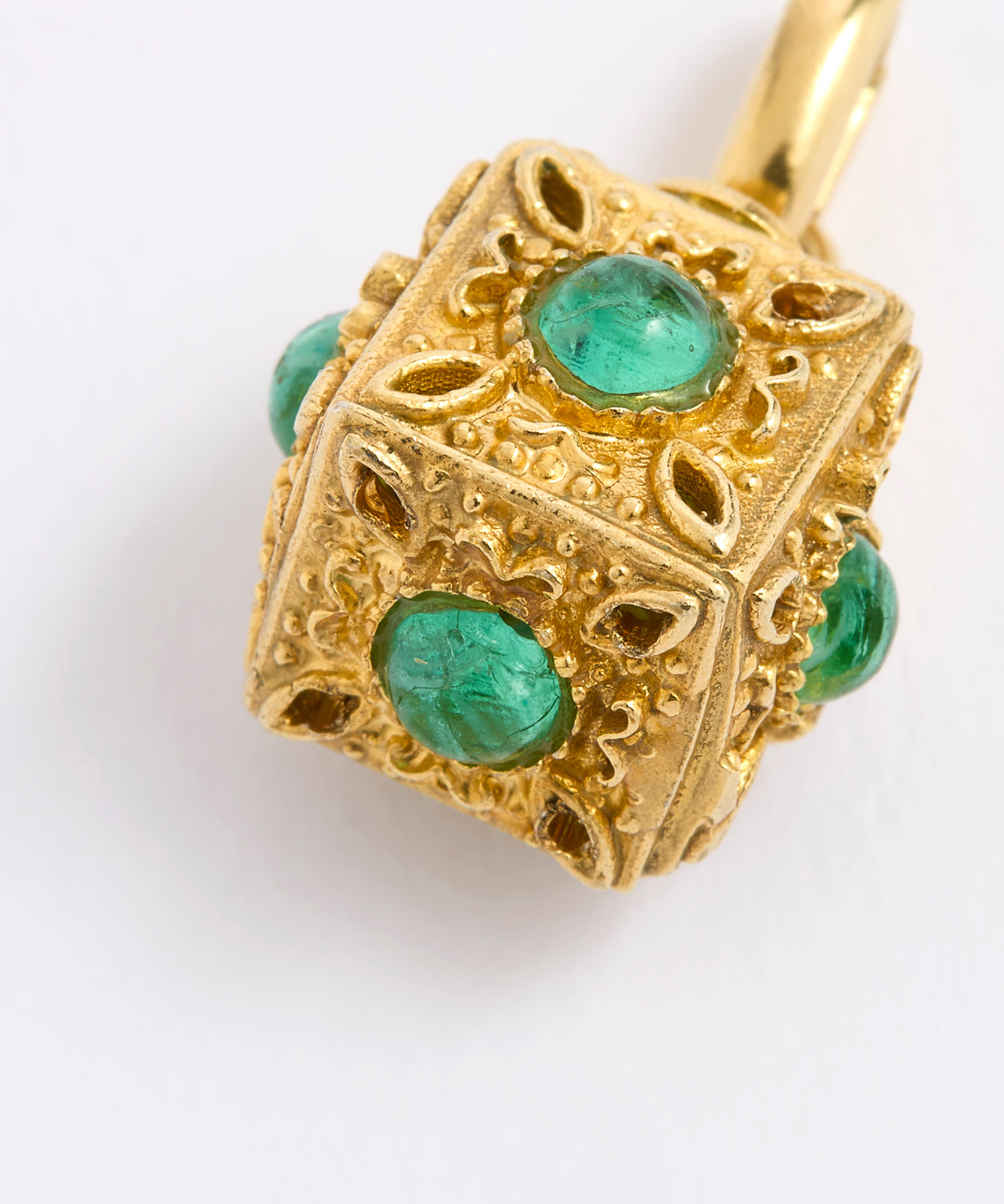 A close-up of a gold pendant shaped like a decorative cube, featuring round, emerald-cut green gemstones set into each side, complemented by an intricate ornate filigree design, displayed against white background.