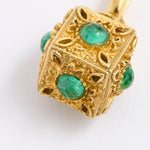 A close-up of a gold pendant shaped like a decorative cube, featuring round, emerald-cut green gemstones set into each side, complemented by an intricate ornate filigree design, displayed against white background.