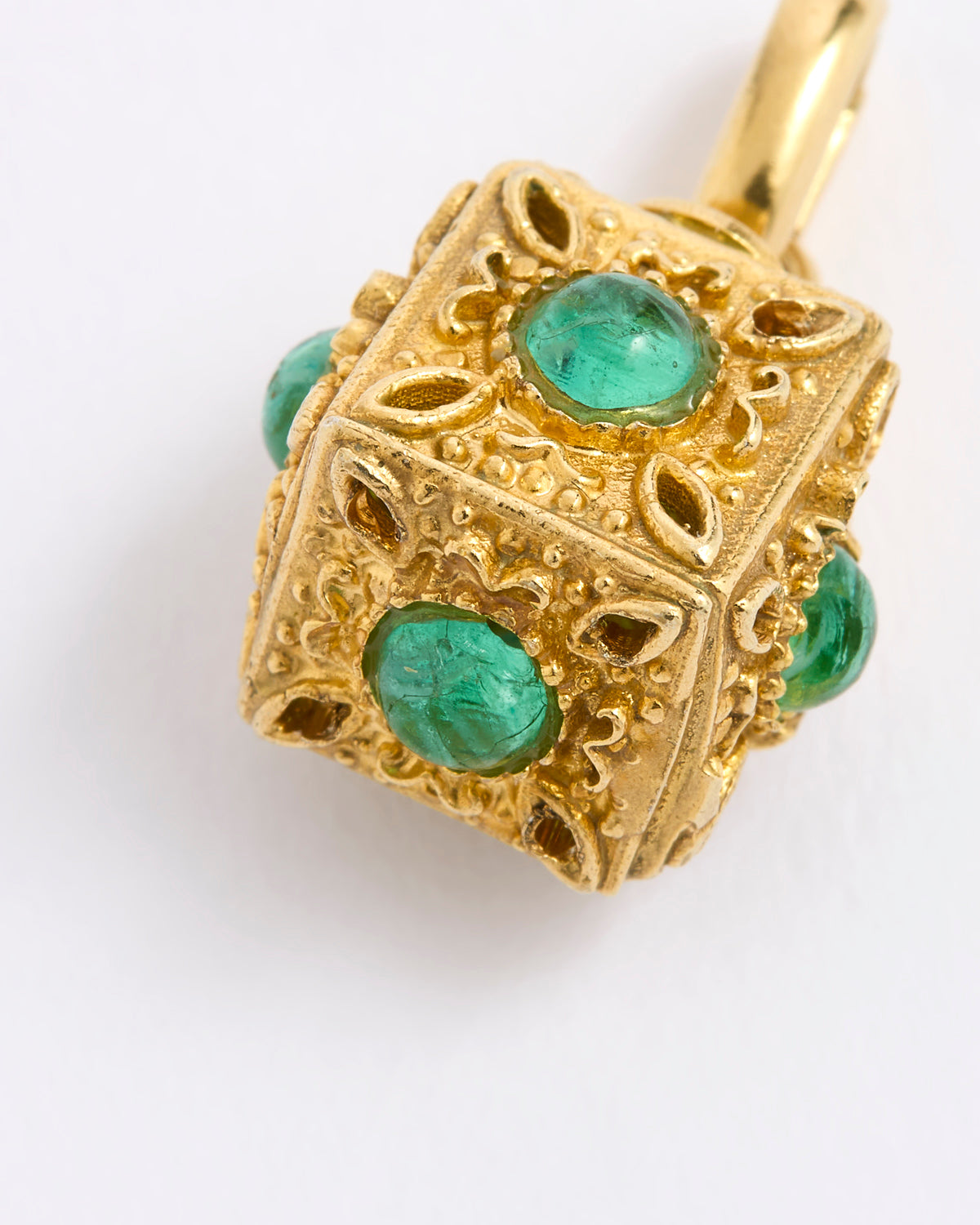 A close-up of a gold pendant shaped like a decorative cube, featuring round, emerald-cut green gemstones set into each side, complemented by an intricate ornate filigree design, displayed against white background.