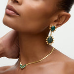 Close-up of a woman wearing gold earrings and necklace with green gemstones on a neutral background