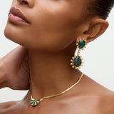 Close-up of a woman wearing gold earrings and necklace with green gemstones on a neutral background