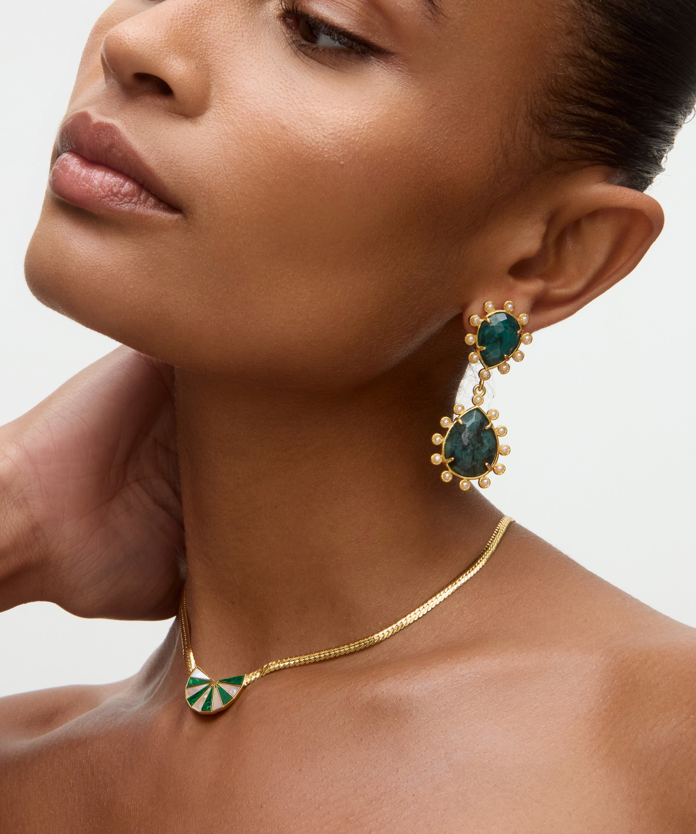 Close-up of a woman wearing gold earrings and necklace with green gemstones on a neutral background
