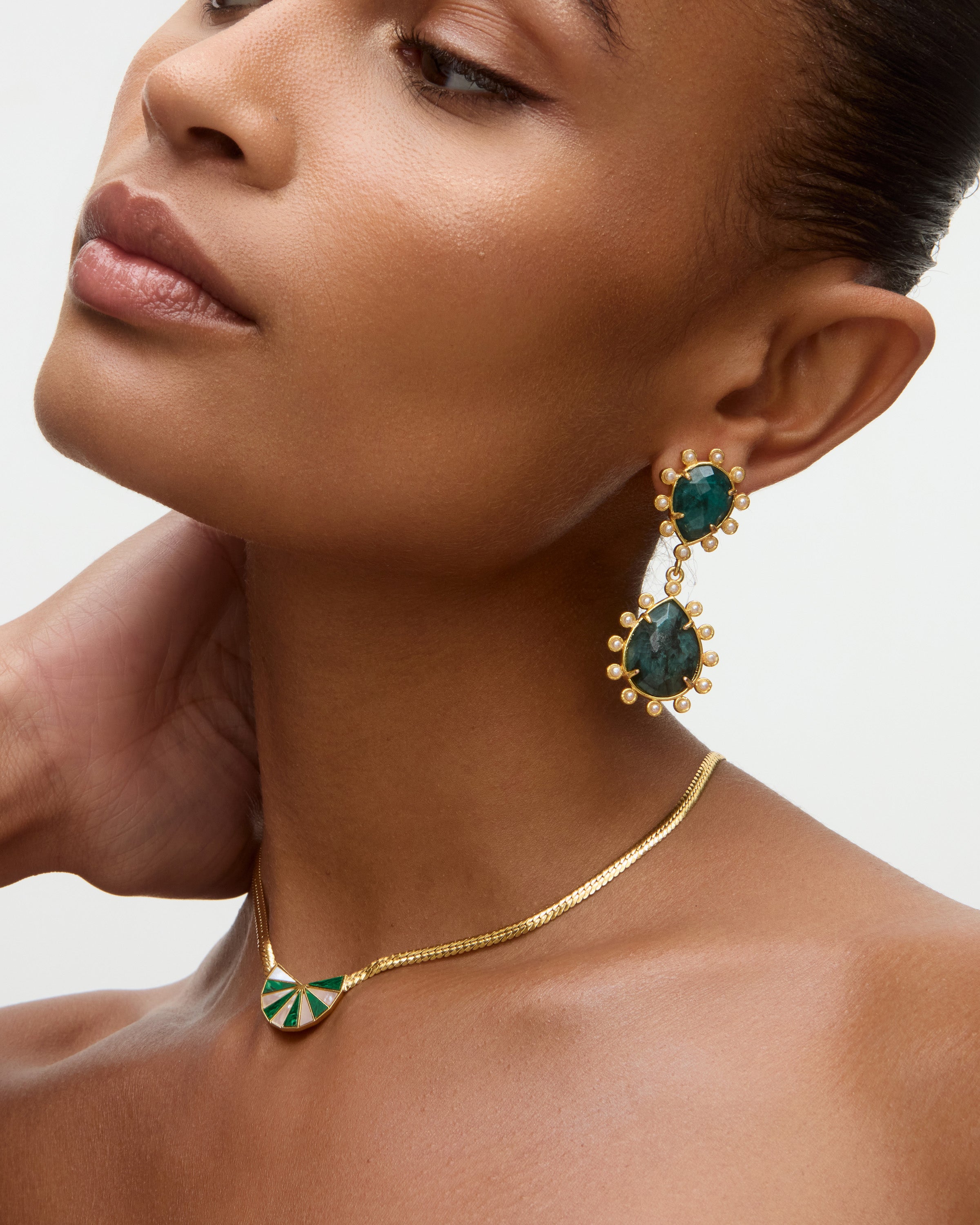 Close-up of a woman wearing gold earrings and necklace with green gemstones on a neutral background