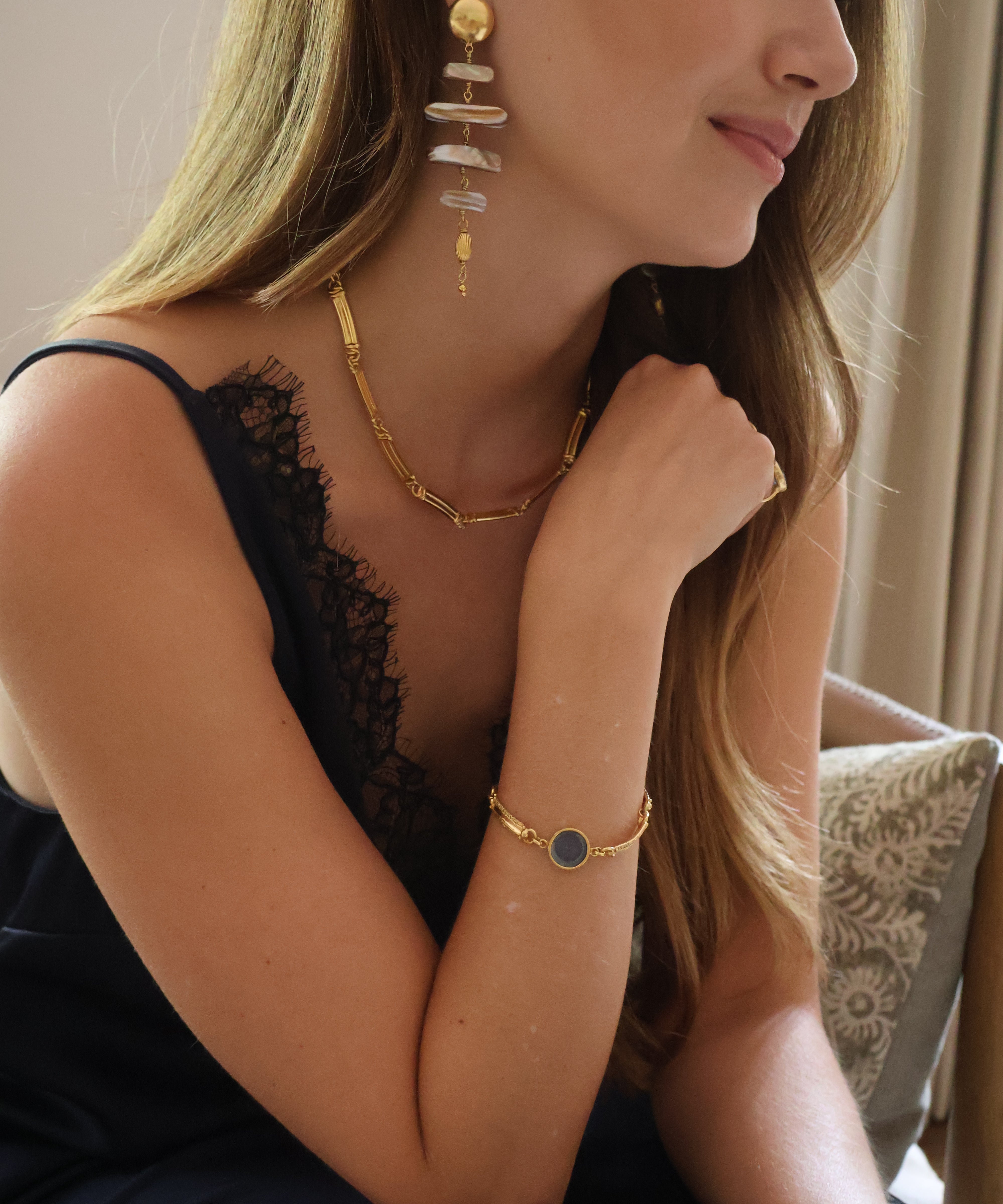 A woman wearing a gold  necklace, matching gold bracelet with a  blue circle center, and a long gold dangle earring with stacked pale stones, posed indoors next to a patterned cushion.