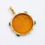 A round gold pendant featuring an amber-colored carved center with green stone accents at the corners, shown on a plain white background.