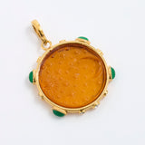 A round gold pendant featuring an amber-colored carved center with green stone accents at the corners, shown on a plain white background.
