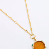 A gold chain necklace featuring a round amber-colored pendant with small green stone accents around its edge, displayed on a clean white background.