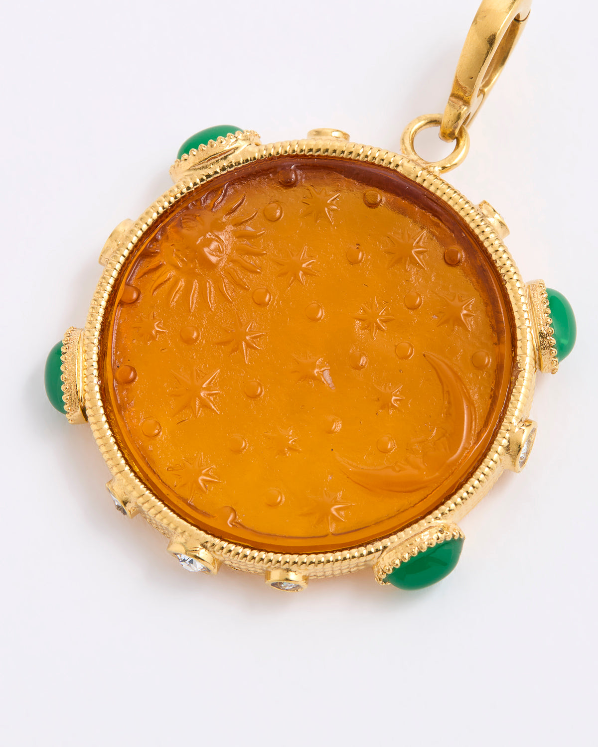 A round amber pendant with a carved sun and moon motif, set in a gold frame with green stone accents, shown on a white background.