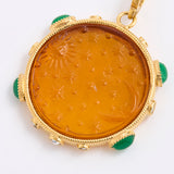 A round amber pendant with a carved sun and moon motif, set in a gold frame with green stone accents, shown on a white background.