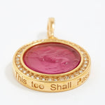 Gold pendant with pink center and engraved text "this too shall pass" on a white background