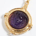 Gold pendant with purple stone featuring a face design on a white background