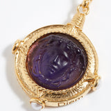 Gold pendant with purple stone featuring a face design on a white background