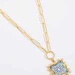 Decorative pendant with blue and gold design on a square tile with mini pearl edging  hung from a chinky gold chain on a white background
