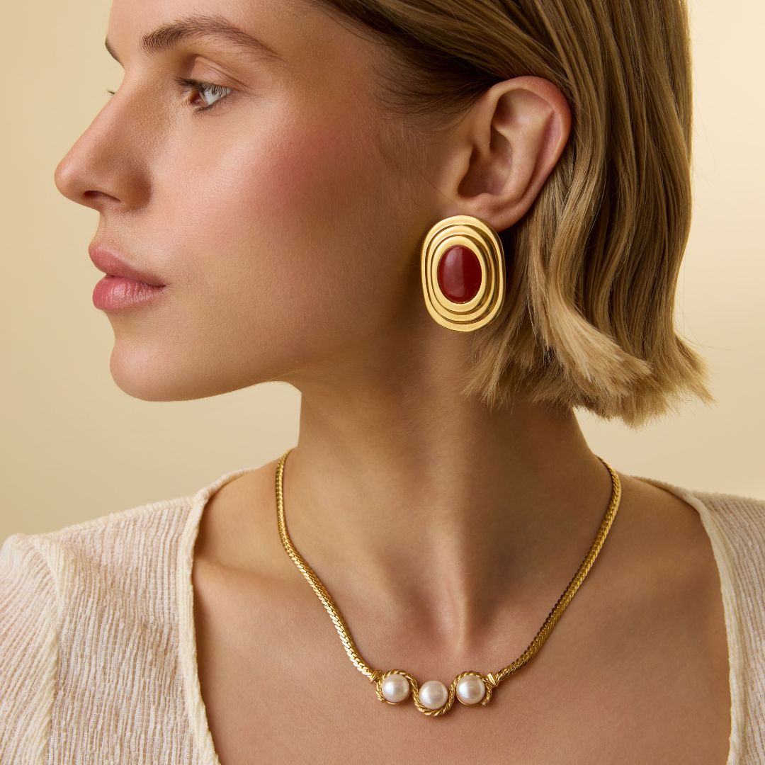 Model shot of lady wearing statement gold earrings with circular top and rectangular drop, with an organic and etched antique finish, on a red background.