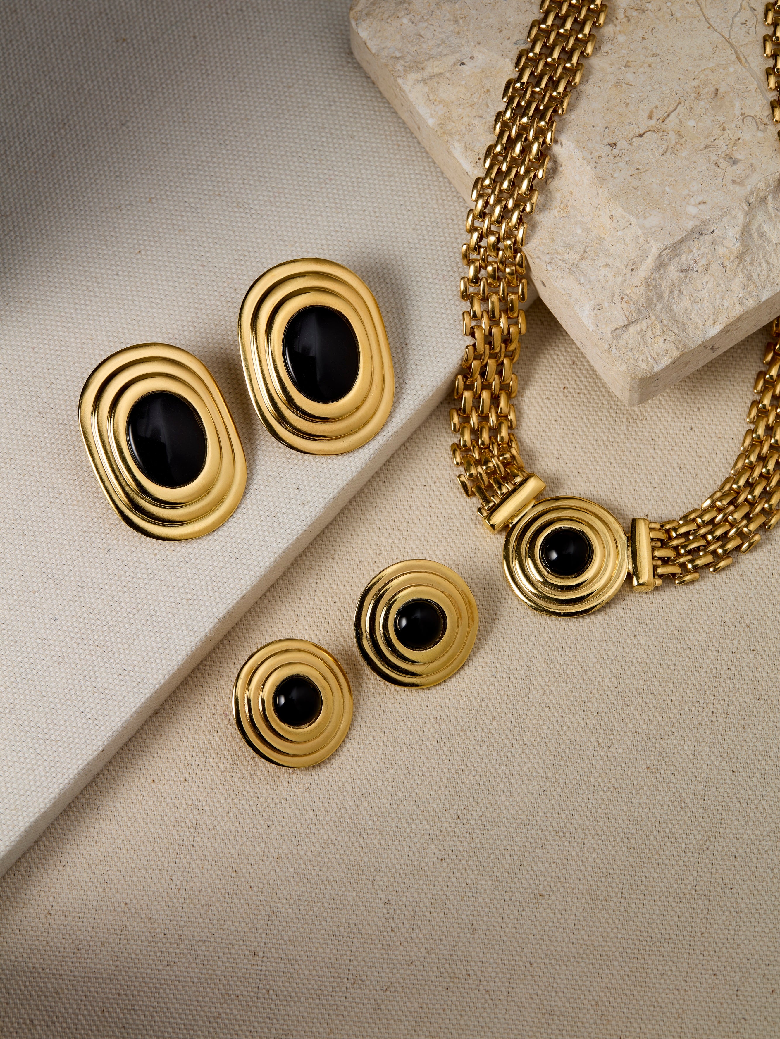 Gold jewellery set with onyx stones on a beige surface, featuring Coco & Roma. 