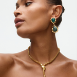 Woman wearing gold snake necklace and emerald green earrings on a neutral background