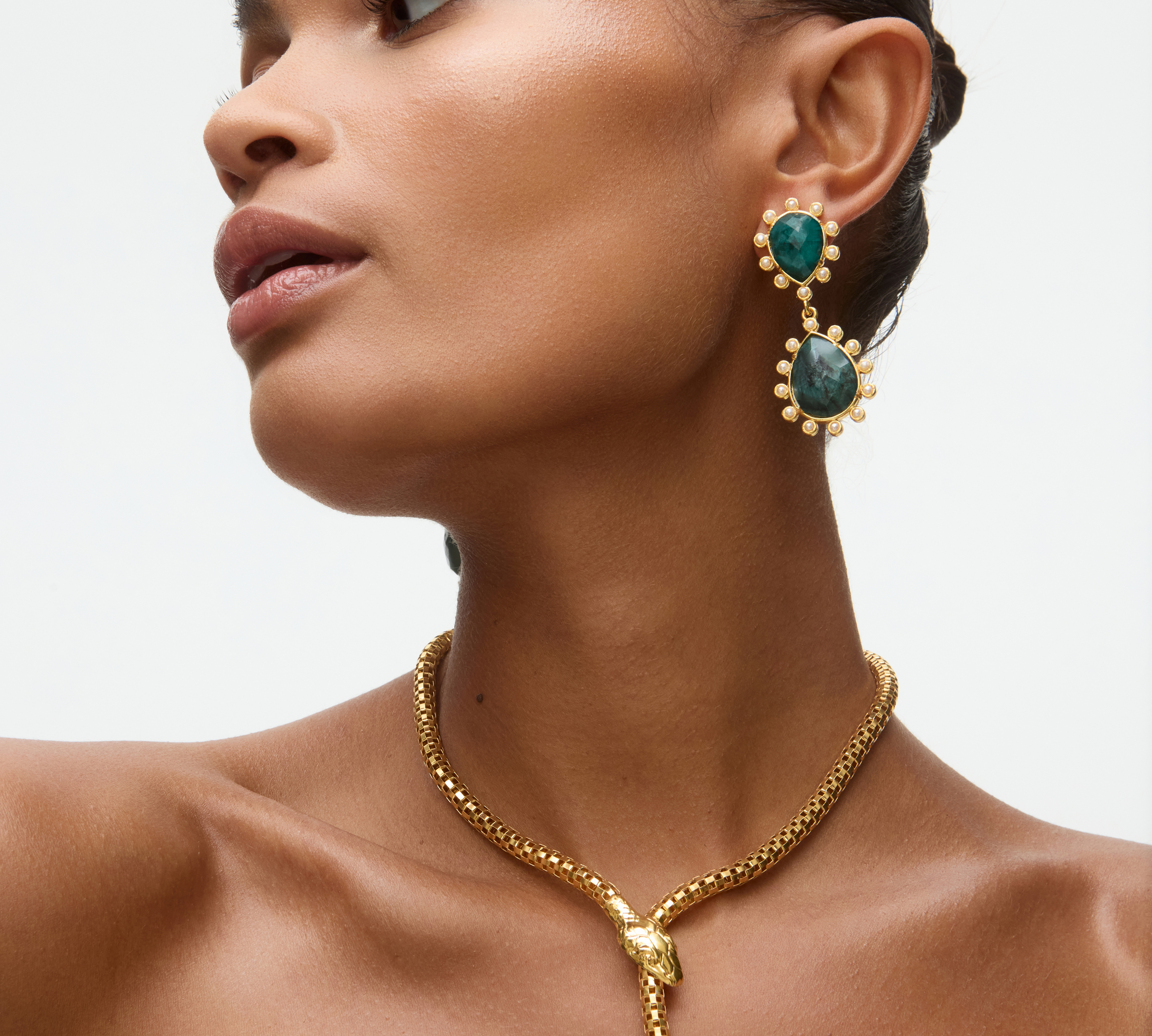 Woman wearing gold snake necklace and emerald green earrings on a neutral background