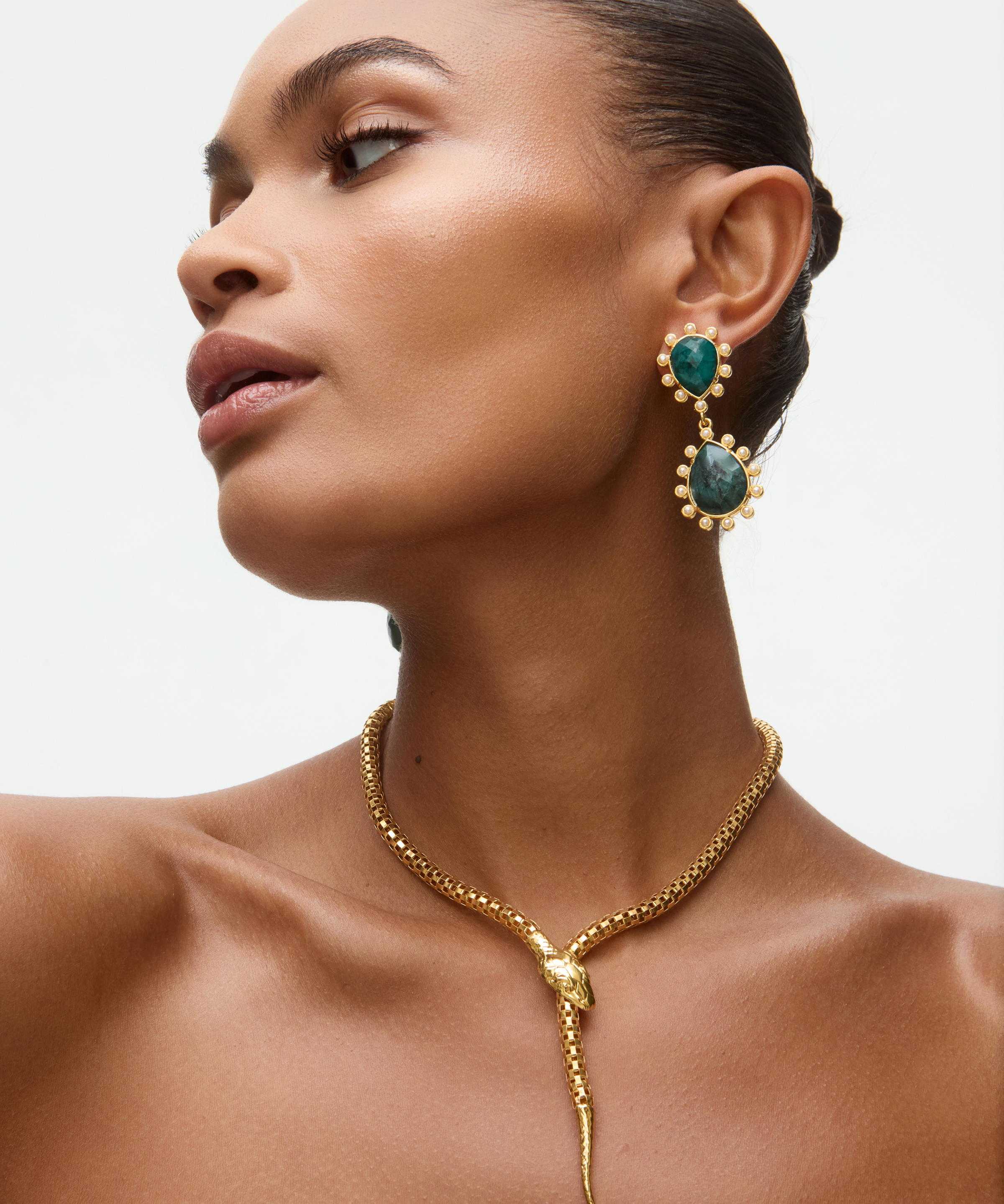 Woman wearing gold snake necklace and emerald green earrings on a neutral background