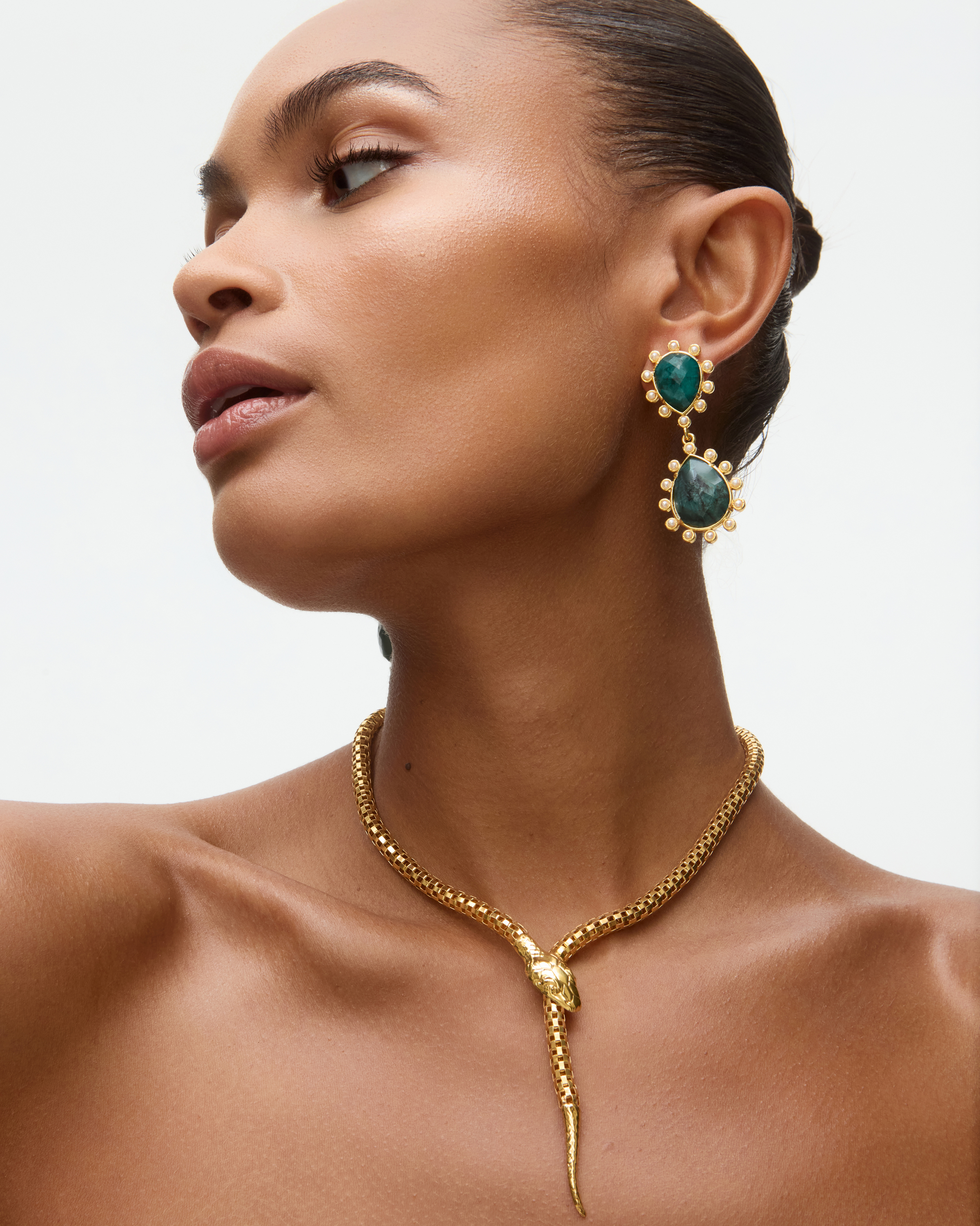 Woman wearing gold snake necklace and emerald green earrings on a neutral background