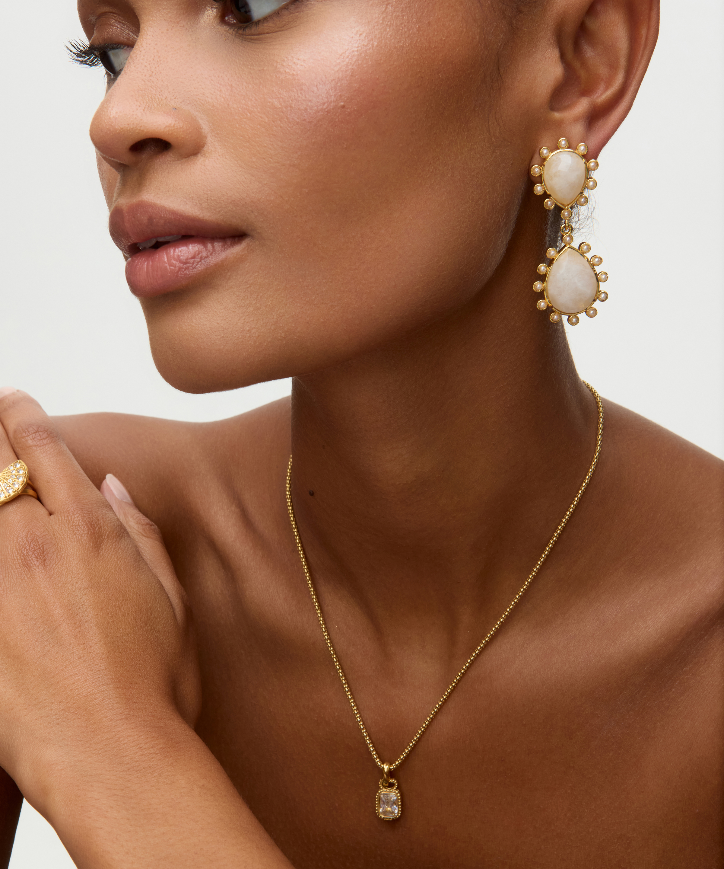 Close-up of a model wearing gold gold earrings with teardrop shaped moonstones and small pearls on  white background earrings and necklace against a neutral background