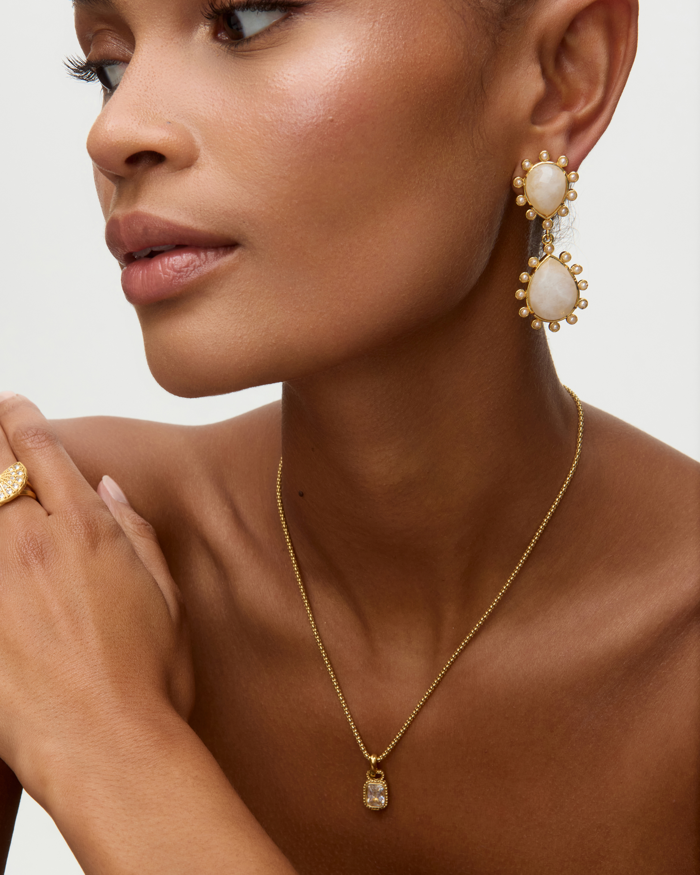 Close-up of a model wearing gold gold earrings with teardrop shaped moonstones and small pearls on  white background earrings and necklace against a neutral background