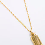 A gold pendant necklace featuring a textured, rectangular handbag-shaped charm with a small gold ring detail above it, suspended from a delicate gold chain on a plain white background.