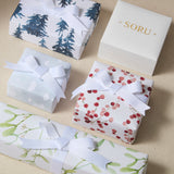 Four wrapped gift boxes with white ribbons are arranged on a beige surface. The boxes feature different holiday-style prints: dark blue pine trees, white clouds, red berries, green leaves, and a plain white box with gold 'SORU' lettering.