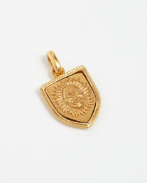 Product shot of a gold plated charm in the shape of a shield with raised engraving of a radiant sun on white background.