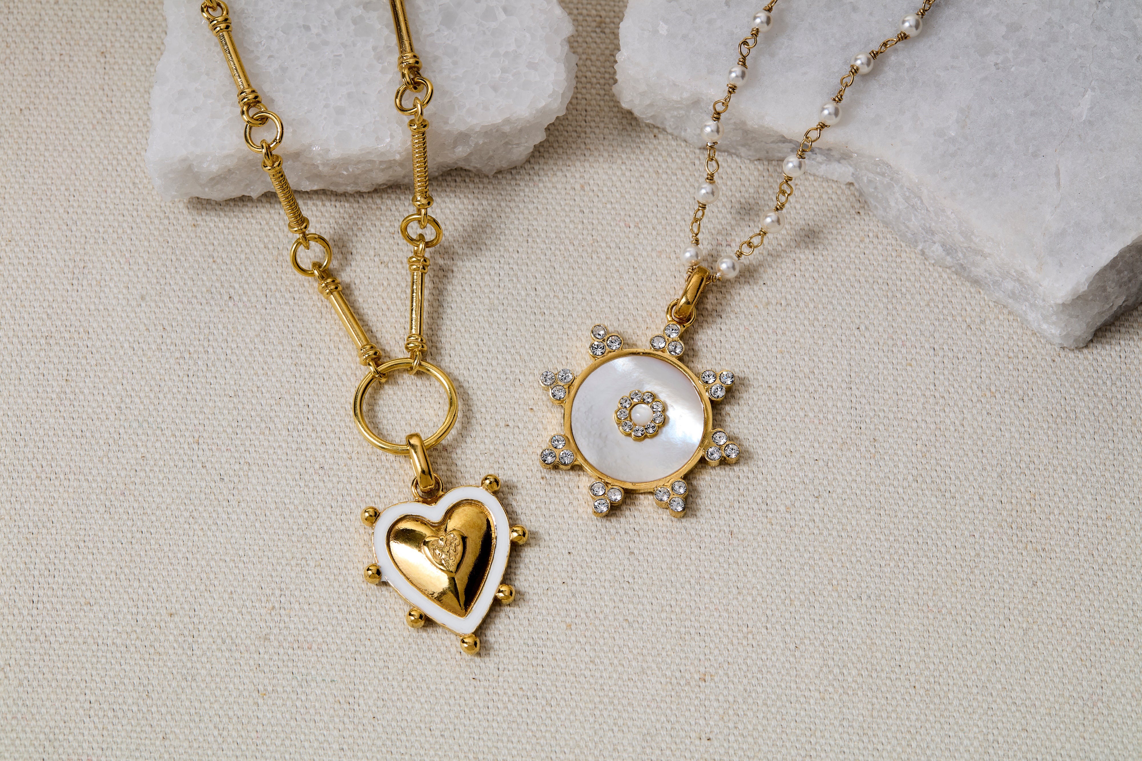 Two gold necklaces with heart and circular pendants on a textured surface.
