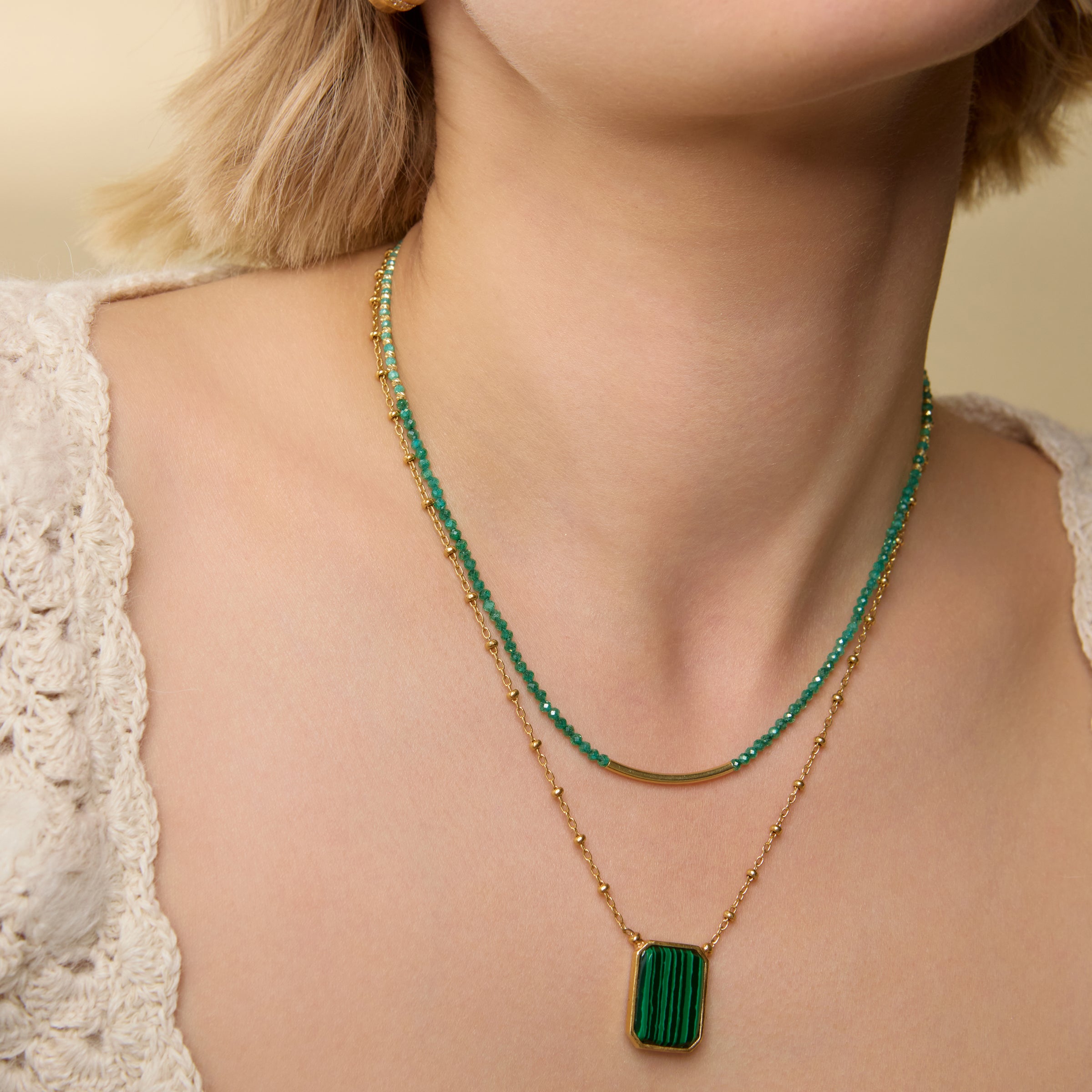 Close-up of a model wearing a necklace featuring rich green agate beads and small gold-toned accents, finished with a polished gold bar at the front centre, layered with another necklace on a neutral background.