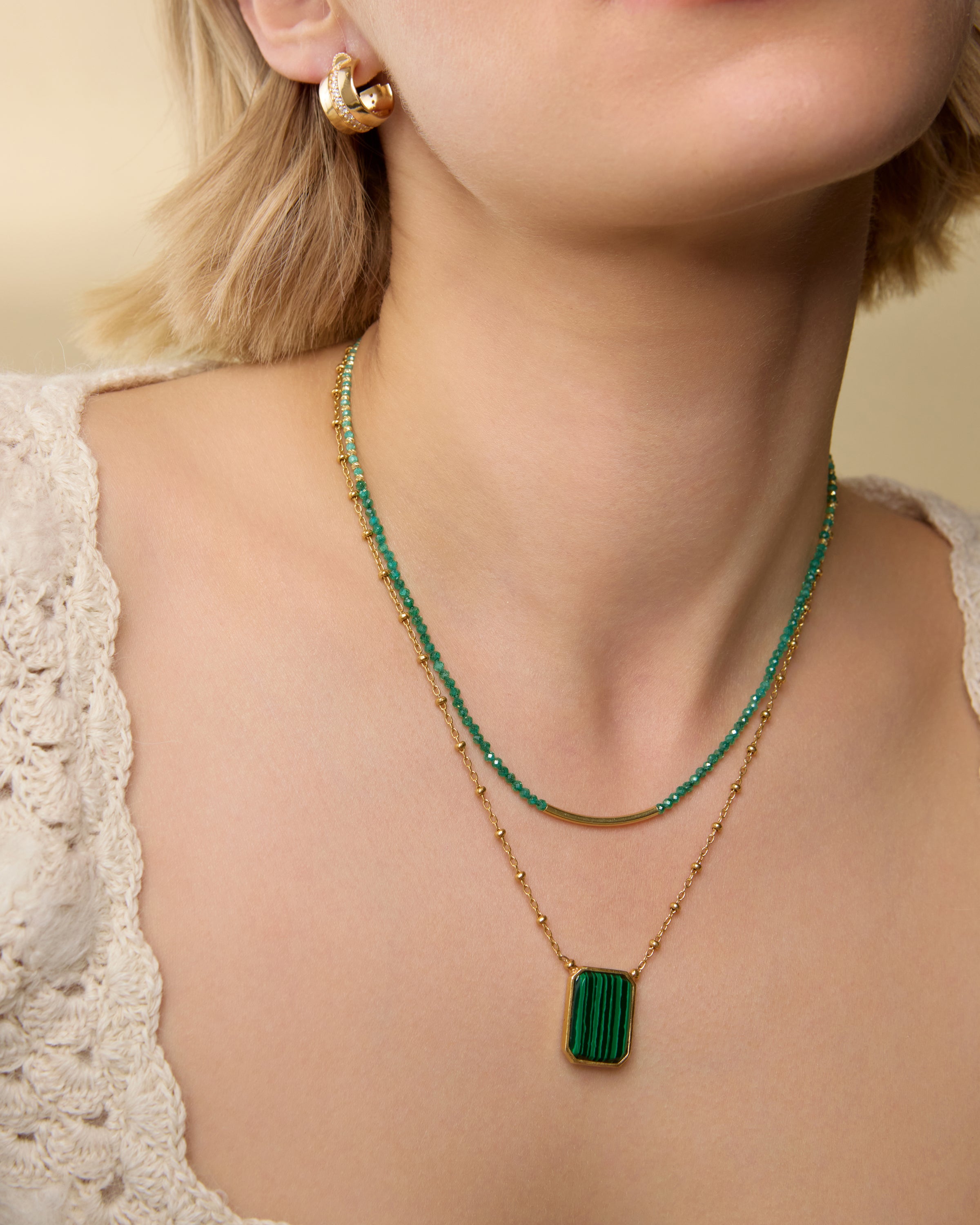 Close-up of a model wearing a necklace featuring rich green agate beads and small gold-toned accents, finished with a polished gold bar at the front centre, layered with another necklace on a neutral background.