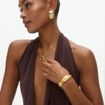 Woman wearing gold jewelry including earrings, necklace, and bracelet on a neutral background