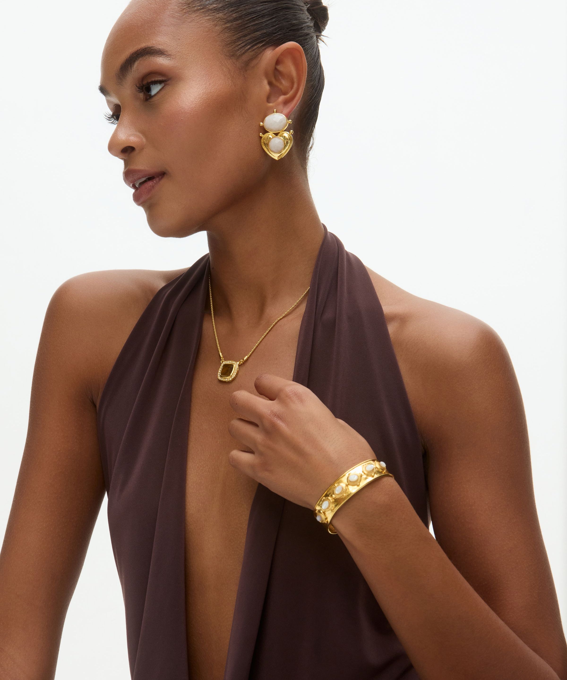 Woman wearing gold jewelry including earrings, necklace, and bracelet on a neutral background