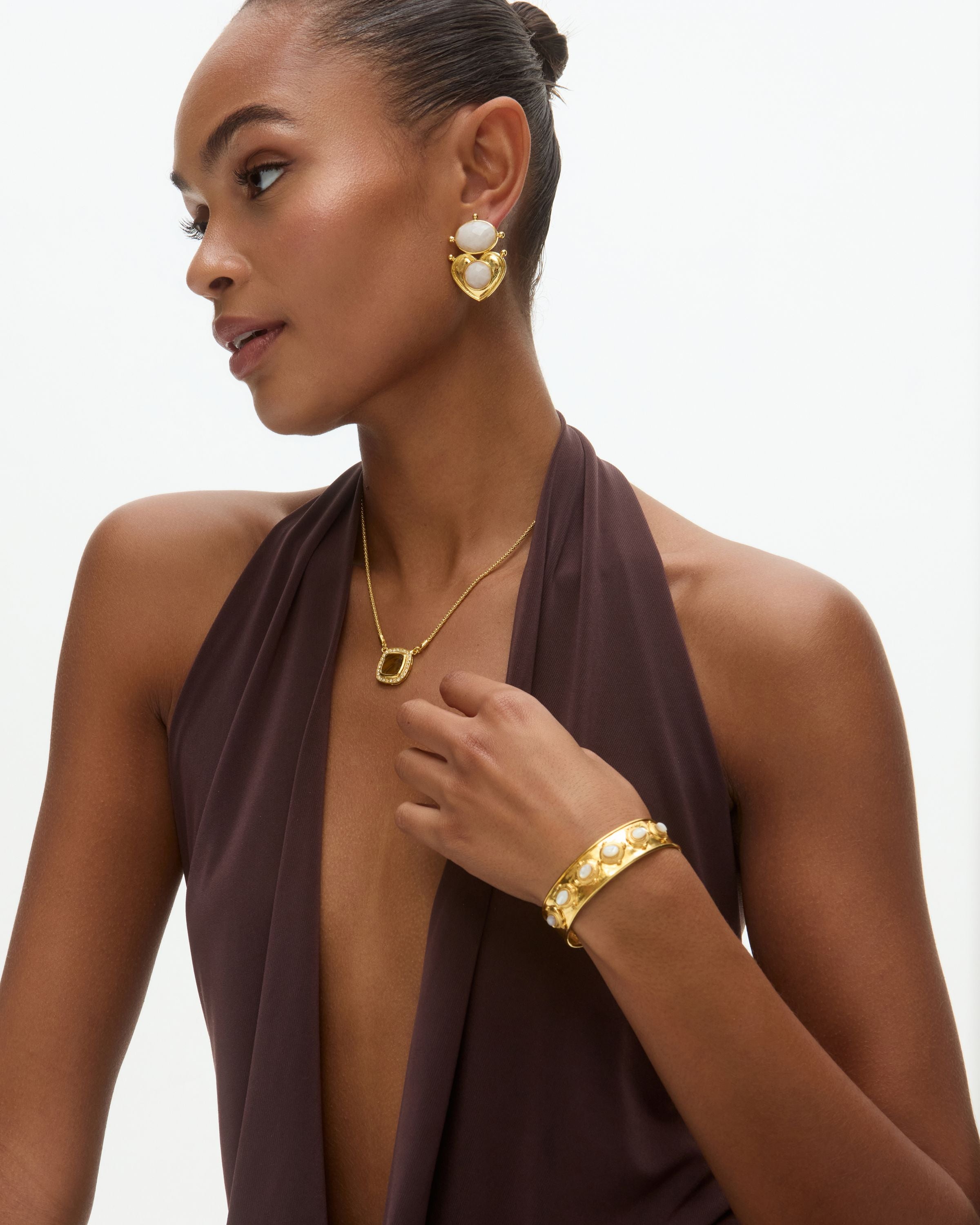 Woman wearing gold jewelry including earrings, necklace, and bracelet on a neutral background