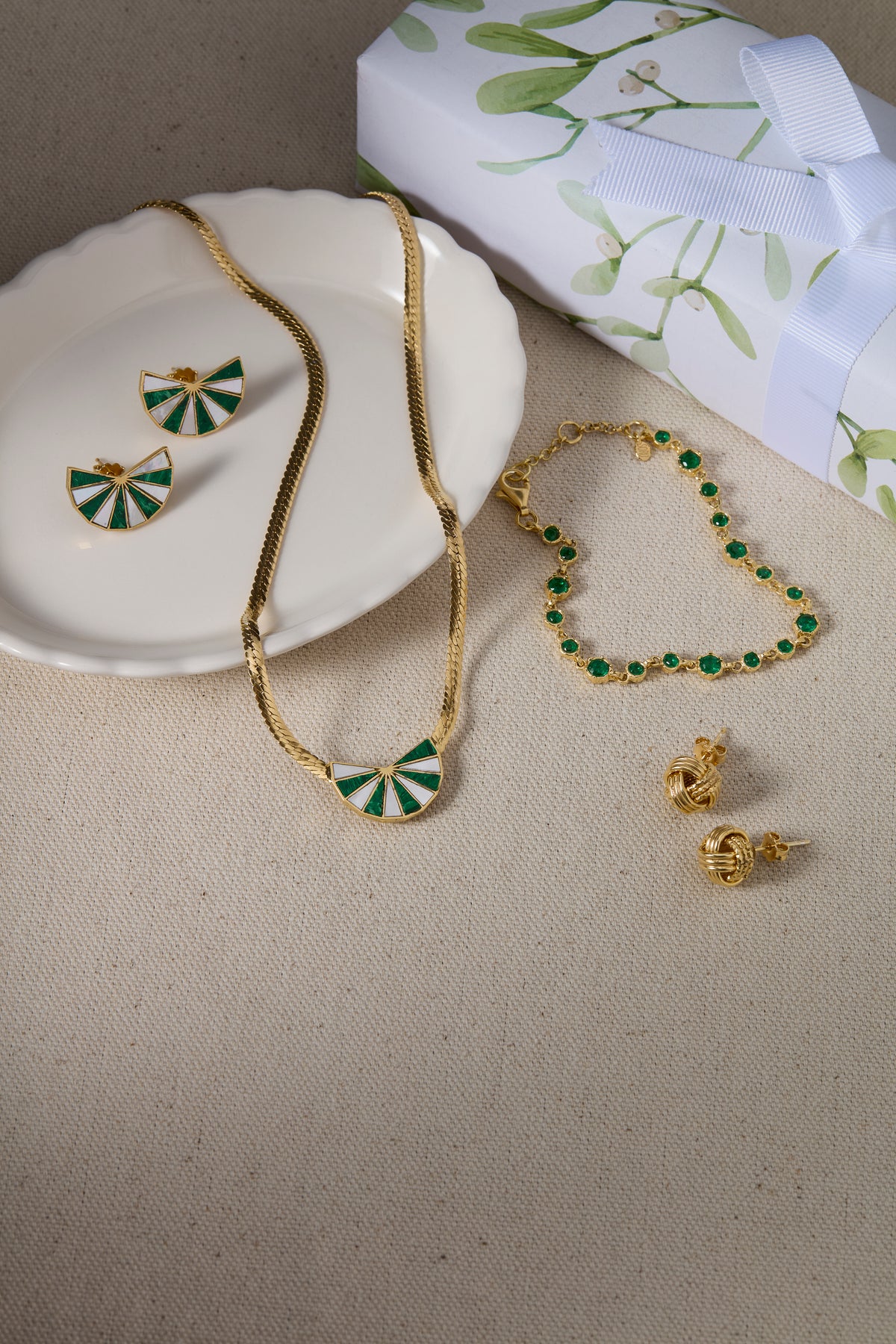 Gold jewelry set with green and white enamel details, including a necklace, bracelet, earrings, and a pair of small gold knotted studs.. arranged on a beige fabric surface next to a white plate and a gift box with a green leaf pattern.