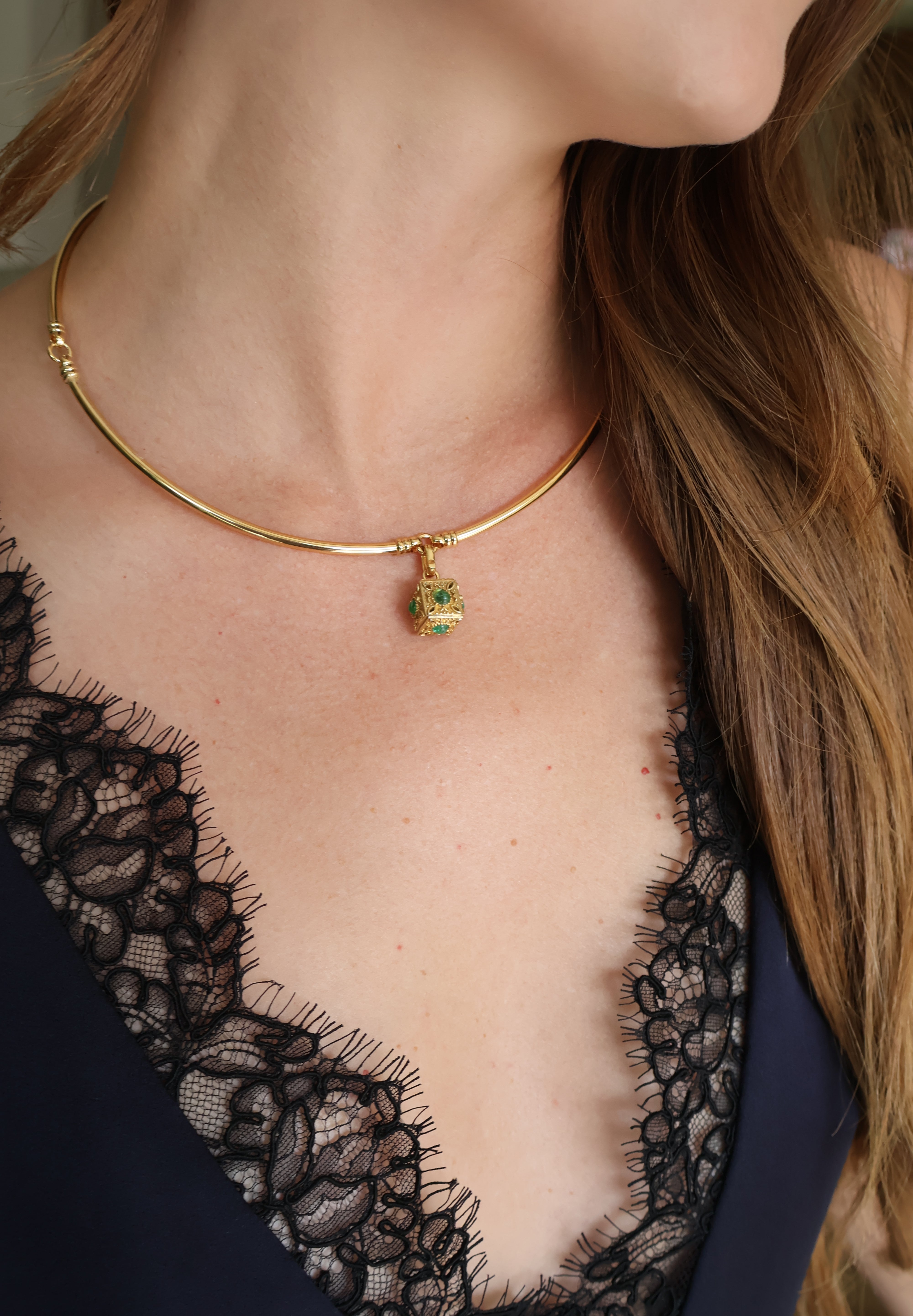 A close-up of a model wearing a delicate gold bangle necklace with a small green  pendant, paired with a black lace top.