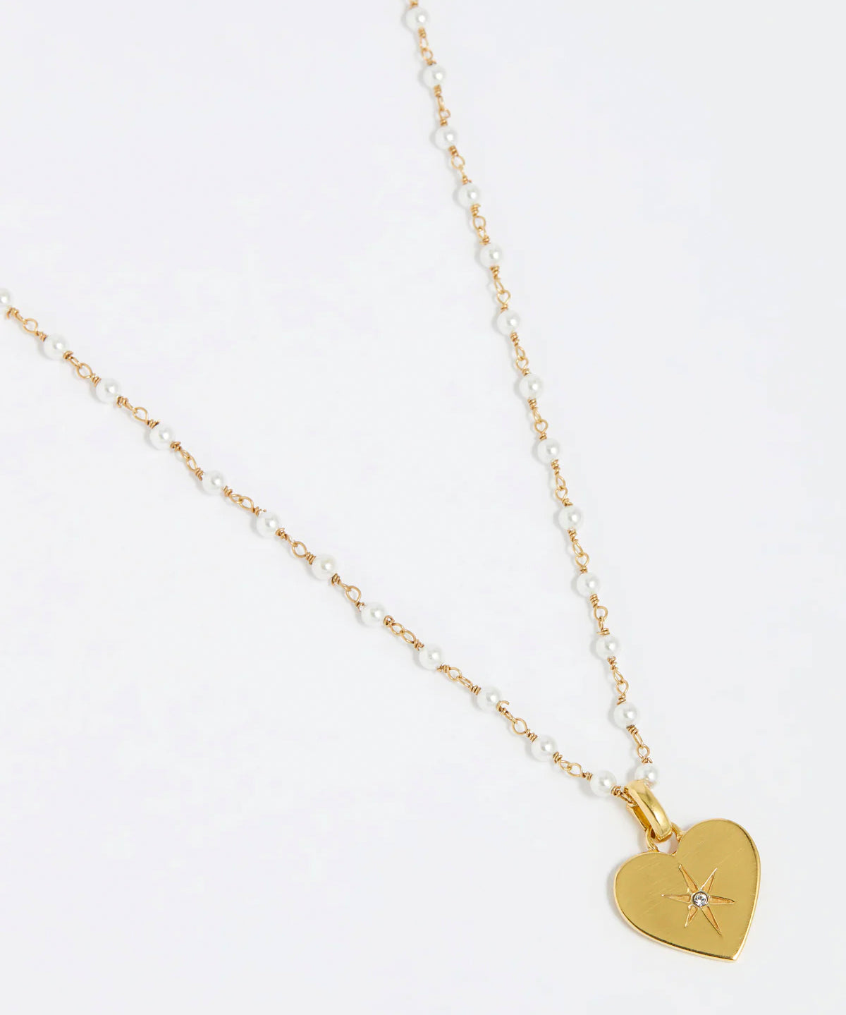 close up image shot of the Soru Jewellery clear crystal surrounded by a star etching set in a shiny gold heart attached to a white beaded necklace on a white background