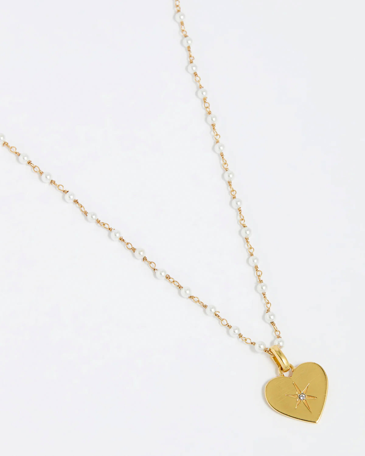 close up image shot of the Soru Jewellery clear crystal surrounded by a star etching set in a shiny gold heart attached to a white beaded necklace on a white background