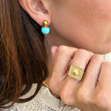 Close-up of a person wearing gold and turquoise earrings and a gold ring with a textured background.