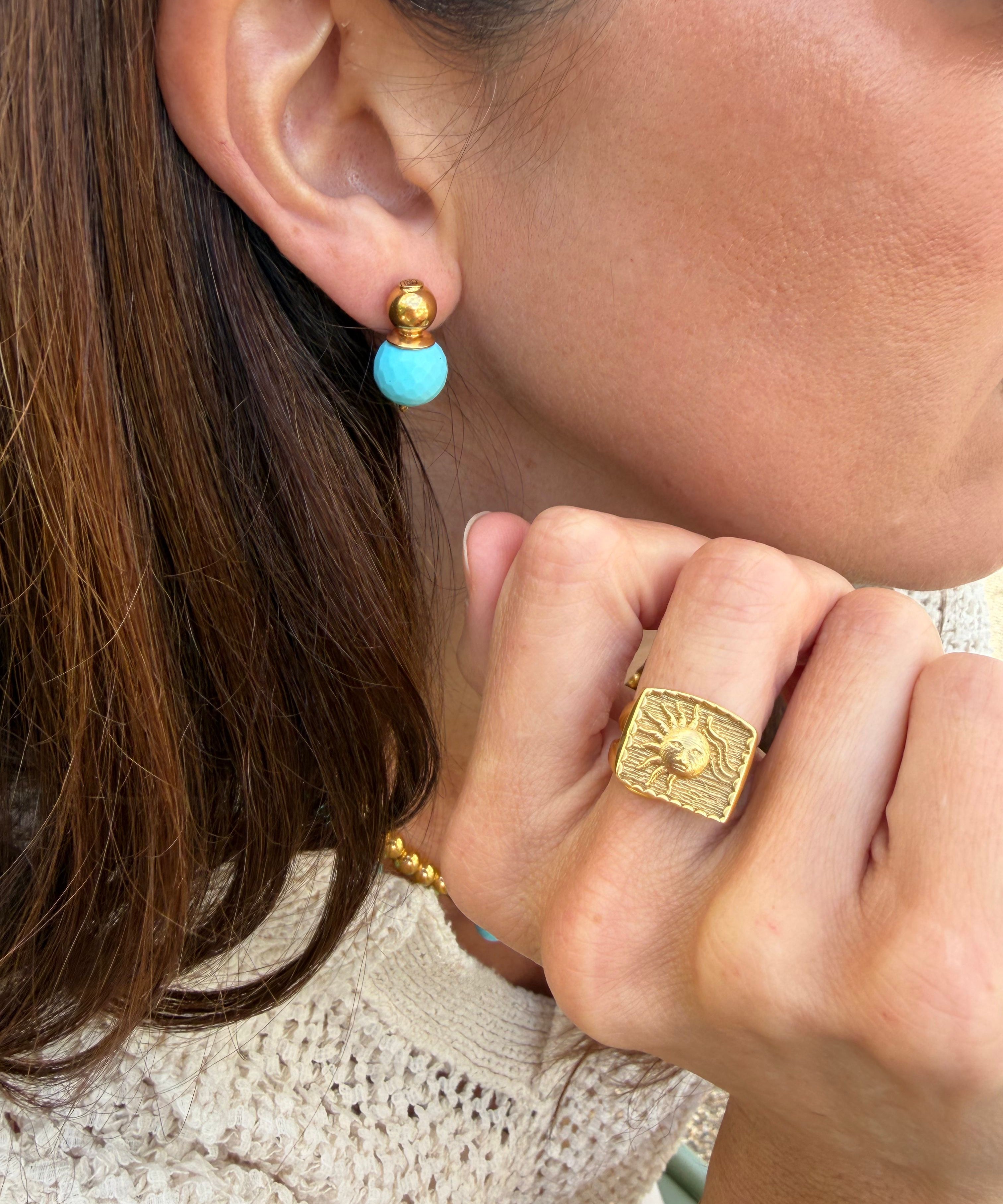 Close-up of a person wearing gold and turquoise earrings and a gold ring with a textured background.